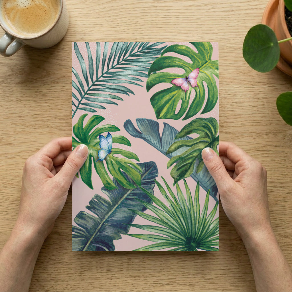 Person holding an art print with tropical leaf design on a wooden table with a cup of coffee and plant.