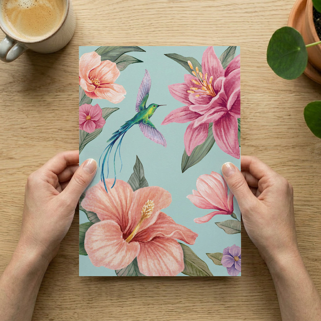 Floral art print with pink flowers and a hummingbird held by hands on a wooden table with a cup of coffee.