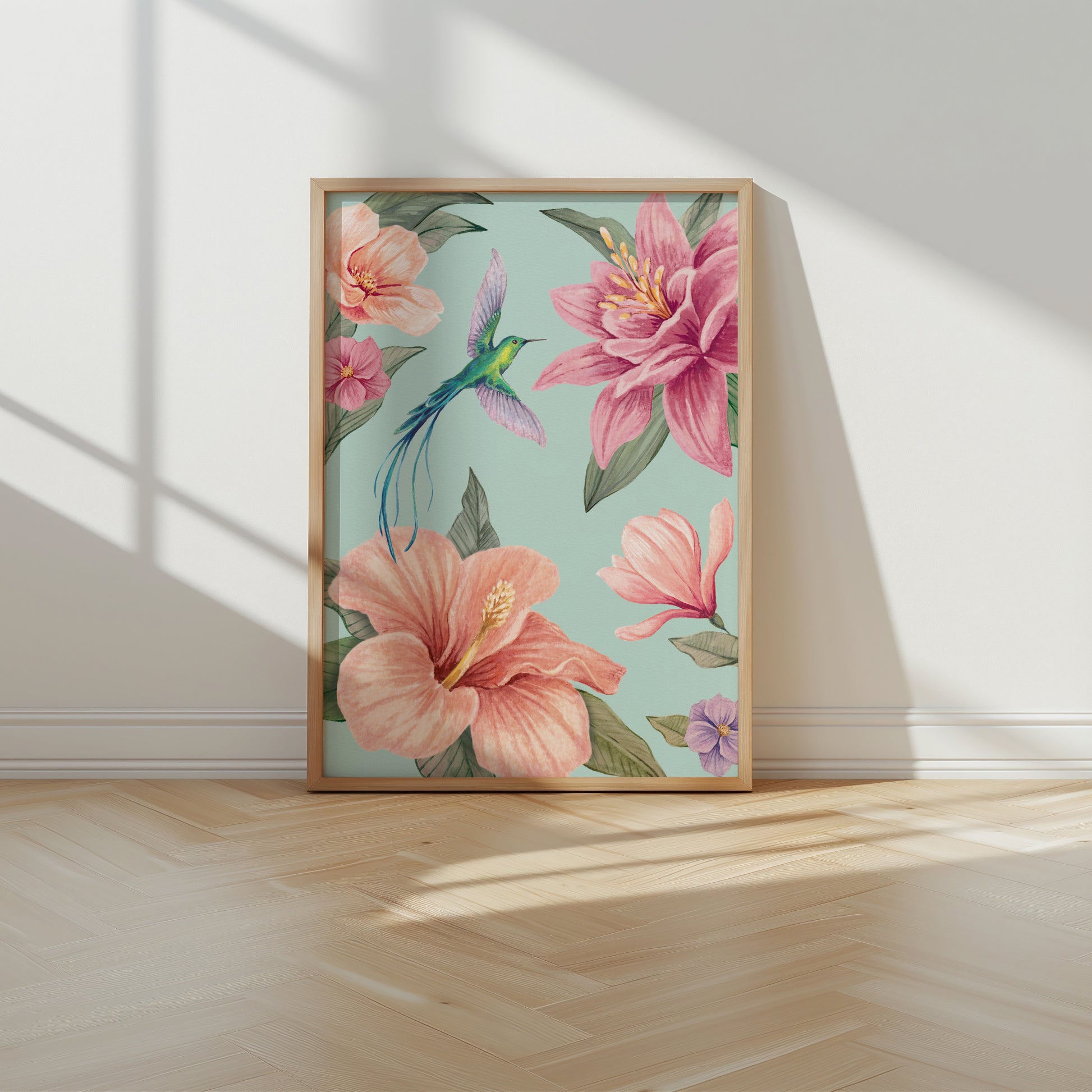 Framed floral artwork with pink flowers and a hummingbird on an aqua blue background, placed on a wooden floor.